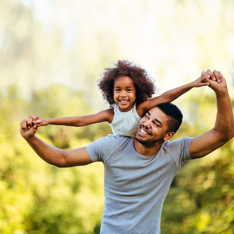 A man in a gray t-shirt holding a young girl s hands above his head in a joyful gesture, both are outdoors with a blurred natural background.